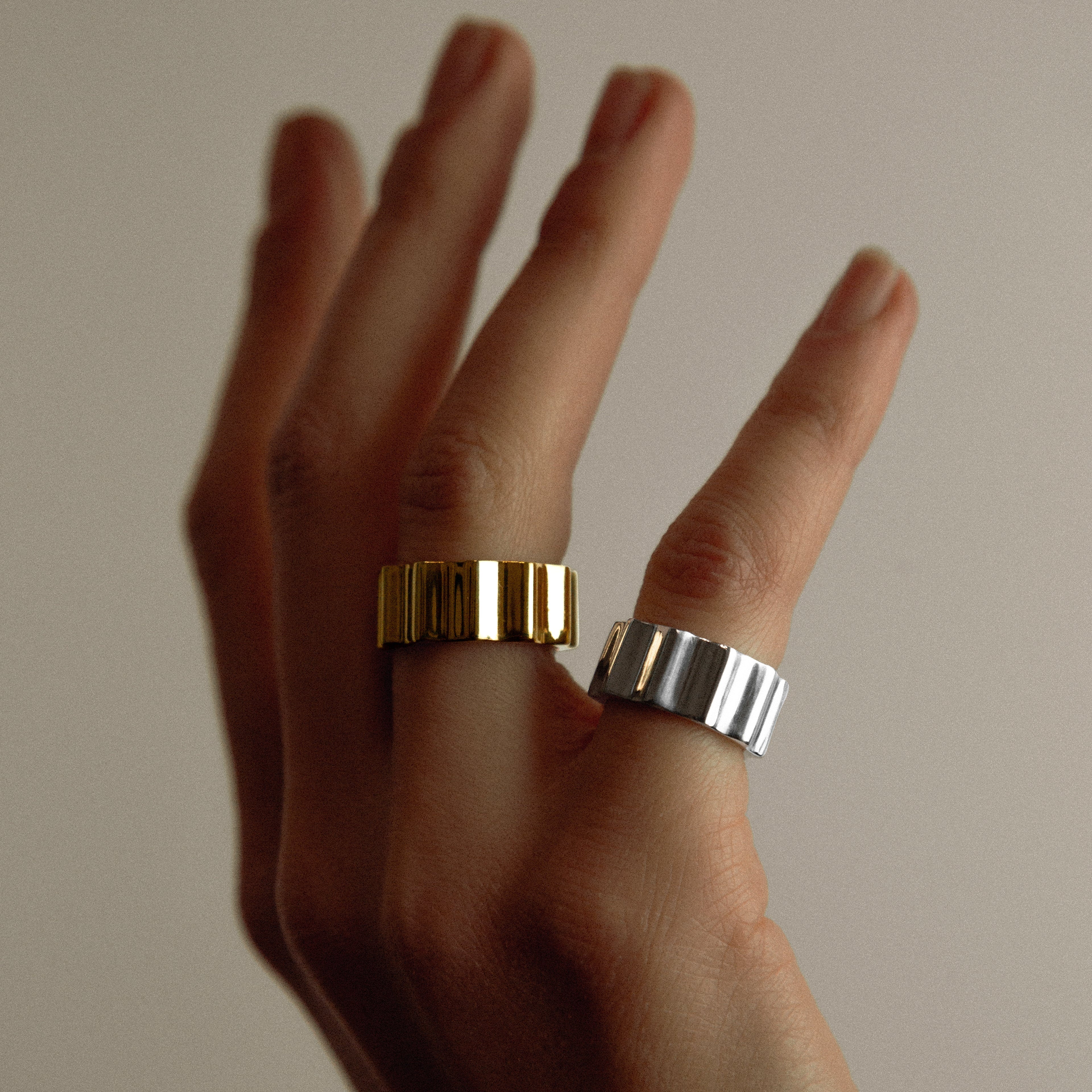Hand wearing two rings, one gold and one silver, against a neutral background