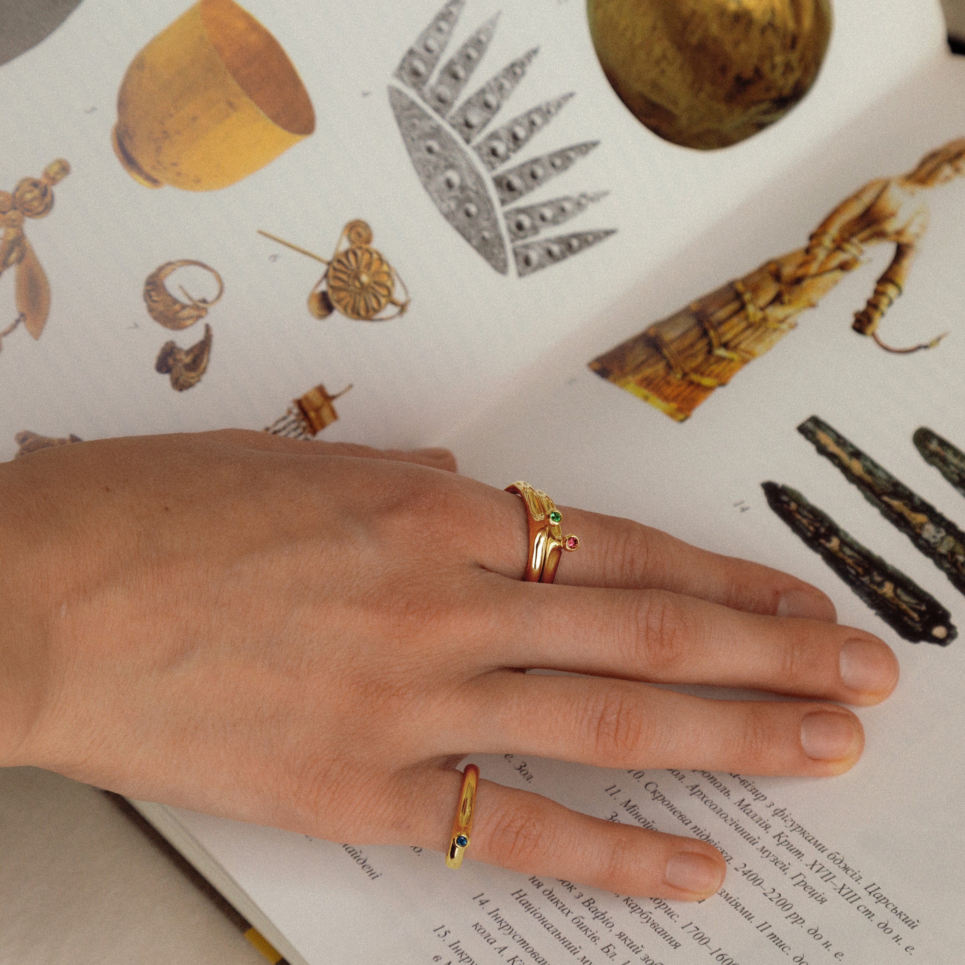A close-up of a person's hand resting on an open book displaying illustrations of Viking-age weapons, including an axe, swords, and other artifacts. The person is wearing two gold rings.